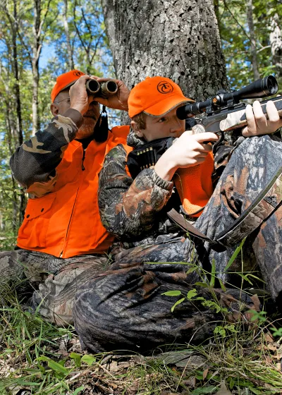 A photo of a young male hunter with his grandfather, both are wearing hunting camo, hunter orange vests, and hunter orange hats. They are sitting at the base of the tree where the boy is aiming his rifle and the grandfather is looking through his binoculars.