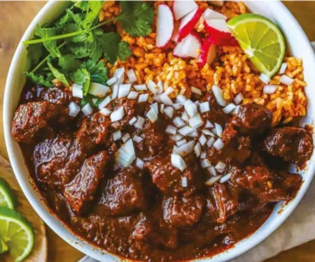 A picture of Chili colorado in a white bowl with a side of rice, garnished with cilatro, radishes, fresh chopped onion, and a slice of lime.