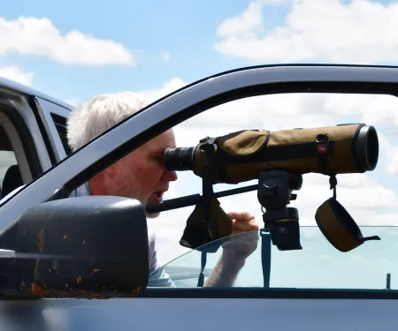 A man looks through a spotting scope attached to an open truck door. 