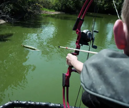 A boy is aiming his red bow at a fish target that is floating on a body of water.