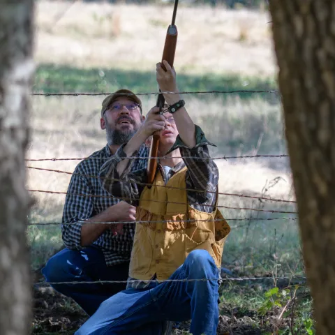 A young man points a gun upward while an adult crouches behind him providing guidance. 