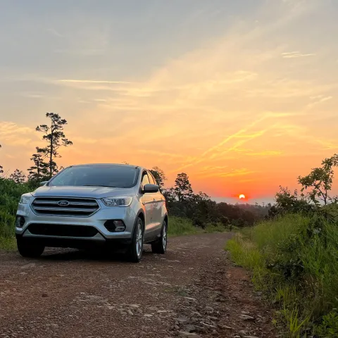A biologist stands in front of a vehicle at daybreak with a data sheet in hand and binoculars and stop watch around his neck. 