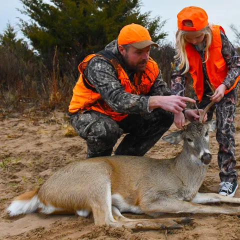 A man and his niece (both wearing hunter orange) are looking at and discussing about the deer that the niece recently harvested.