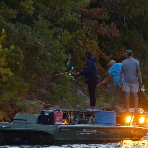 Two men and a woman stand on a boat with bows and arrows in hand. 