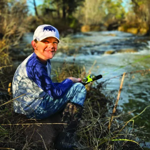 A young man sits at the edge of a waterway with a fishing pole in hand. 
