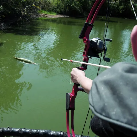 A boy is aiming his red bow at a fish target that is floating on a body of water.