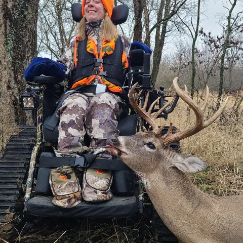 Hunter wearing orange pictured with a harvested deer. Note from her father, Mike Burnside - "My daughter Kenzi uses a vacuum air actuator to fire the rifle. She sucks on a vacuum tube, the vacuum is converted to electric signal via attached 12v battery, and the cables go to a trigger guard mount, which pushes actuator against the trigger."