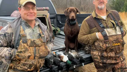 Two men stand next to a truck with a dog and five ducks on a tailgate. 