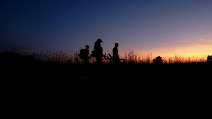 A group of archery hunters are walking through a field at sunrise.
