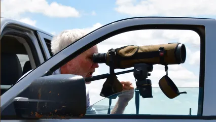 A man looks through a spotting scope attached to an open truck door. 