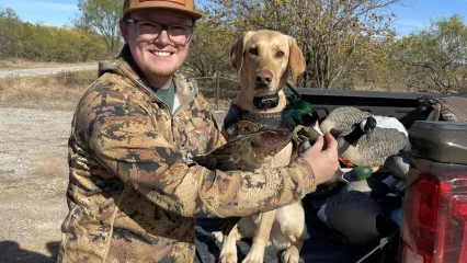 A man holds a duck with a brown body, green head, and white-ish bill in front of a yellow dog that is sitting on a truck tailgate. 