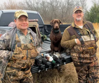 Two men stand next to a truck with a dog and five ducks on a tailgate. 