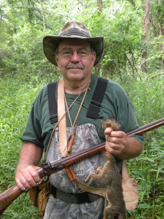 A man in a hat holds a gun in one hand and a squirrel in the other. 