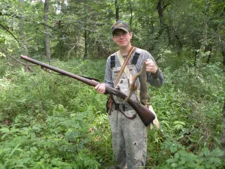A young man stands in a wooded area with a gun in one hand and a squirrel in the other.