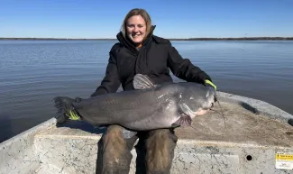 A woman in a black coat holds a large fish on her lap. 