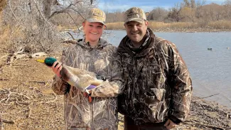 A youth hunter is standing next her dad while smiling and holding her harvested duck.