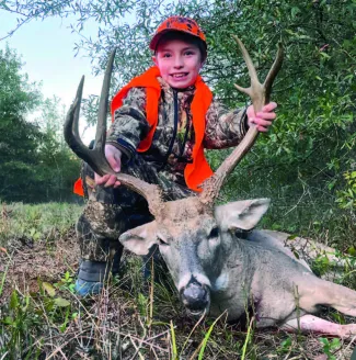 A boy in hunter orange is holding the the antlers of his recently harvested white-tailed buck.