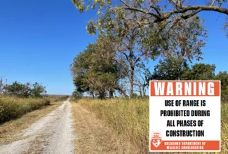 A gravel road runs through tall grass and trees under a clear blue sky. A large warning sign in the foreground reads, "WARNING: Use of range is prohibited during all phases of construction" with the Oklahoma Department of Wildlife Conservation logo.
