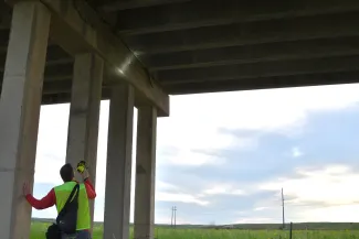 A man in a reflective vest shines a light on the underside of a bridge. 