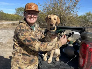 A man holds a duck with a brown body, green head, and white-ish bill in front of a yellow dog that is sitting on a truck tailgate. 