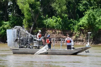 Three people navigate a river on an airboat with nets hanging over the sides of the boat. 