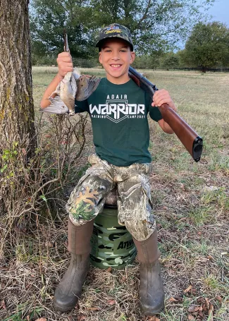 A smiling boy poses with a shotgun over a shoulder and his harvested dove in his hand.