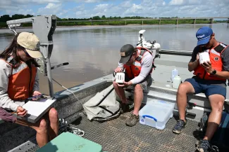 Three people sit on the deck of a boat. One person holds a clipboard and the other two are looking into small white containers.
