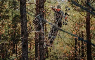 A bowhunter is in a sling tree stand while aiming his bow downward.