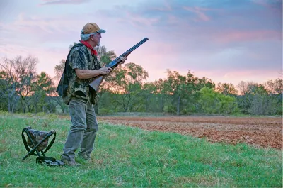 A dove hunter stands at an open field with a stool behind him. 